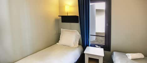 Una camera d'albergo piccola con due letti singoli, un comodino, una lampada e una finestra con tende.