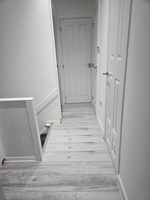 A narrow hallway with wooden flooring and white walls, leading to a closed door at the end.