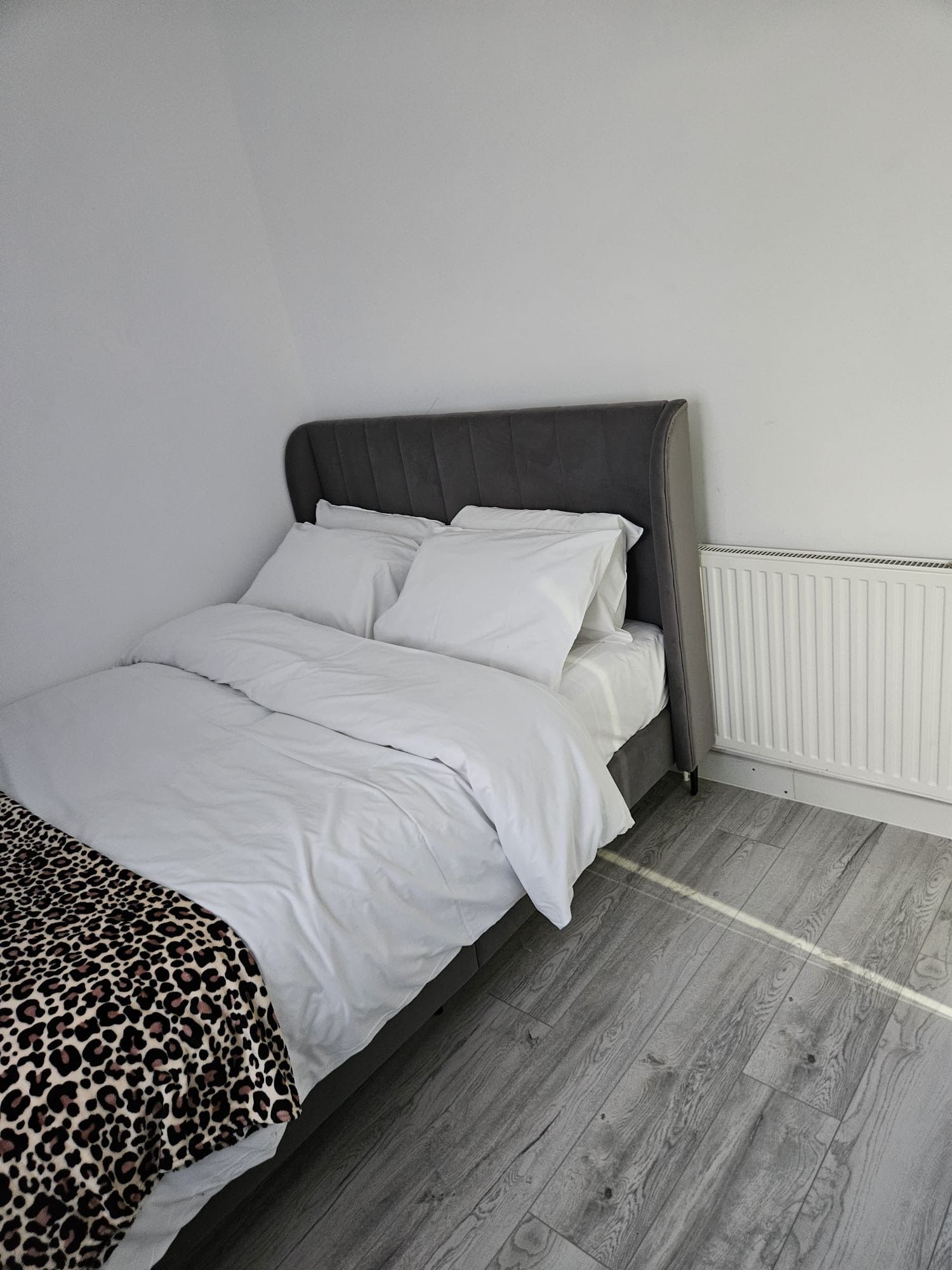 Una cama con ropa blanca y un cabecero gris en una habitación con suelo de madera y un radiador blanco.