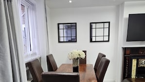 A dining area with a wooden table and six chairs, a vase of flowers, and a fireplace with a television. - Spacious 4-Bed House Near Manchester and Trafford Centre, Free Parking & WiFi (Partington)