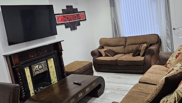 A modern living room with a flat-screen TV mounted on the wall, a brown sofa, a wooden coffee table, and a fireplace with decorative items. - Spacious 4-Bed House Near Manchester and Trafford Centre, Free Parking & WiFi (Partington)