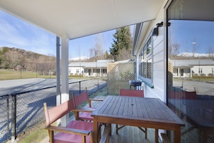 Un patio exterior con mesa y sillas de madera, un área cubierta con vista a un edificio y una montaña al fondo.