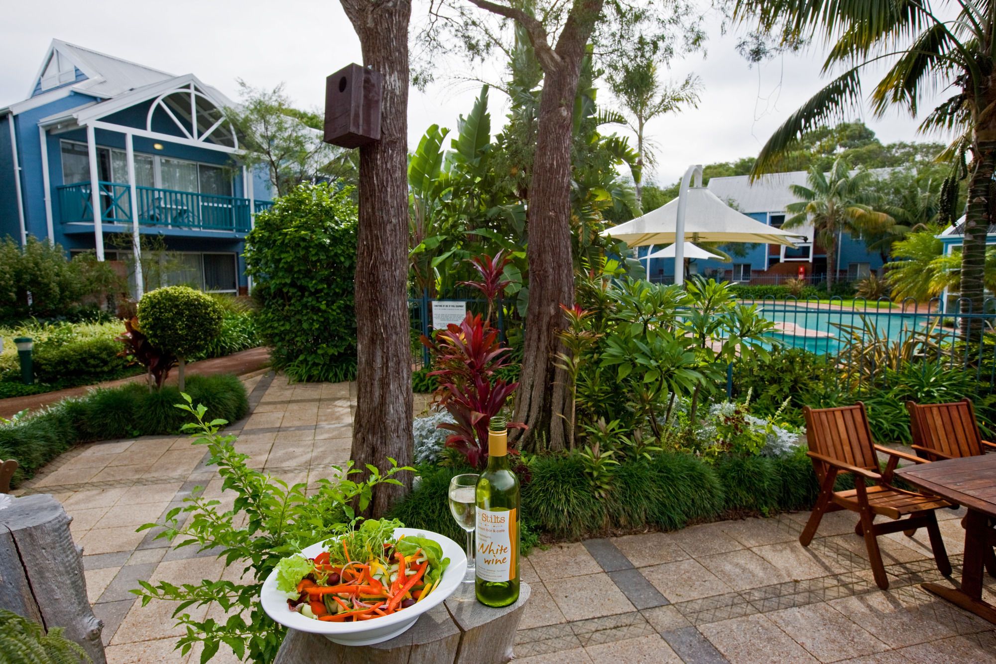 Un área de comedor al aire libre con una mesa y sillas, una piscina al fondo y diversas plantas y árboles.