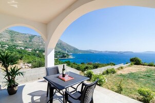 A terrace with a table and chairs, overlooking a coastal landscape with hills and the sea.