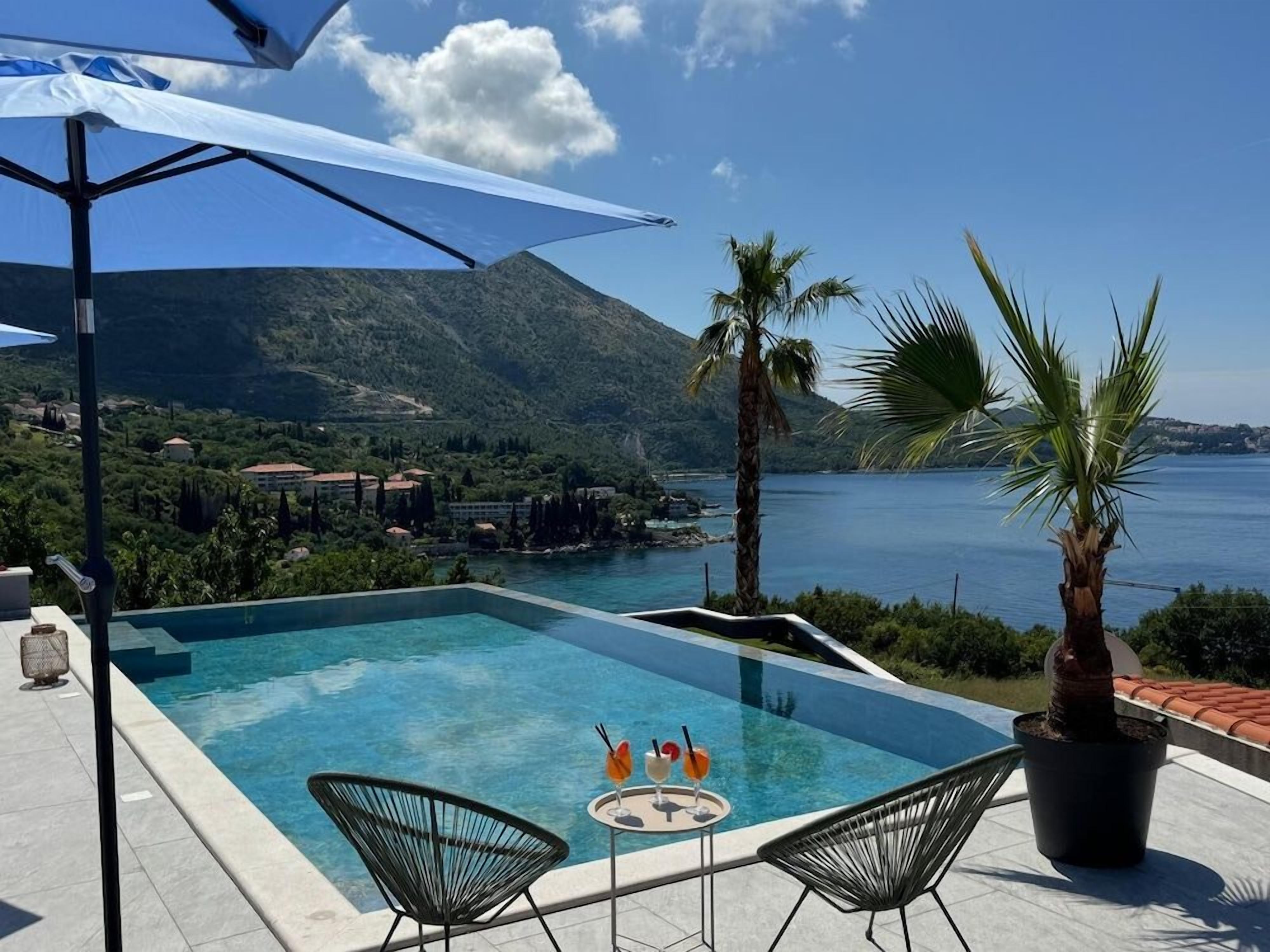 Una piscina infinita con vistas a una montaña y al mar, una mesa con bebidas y dos sillas.