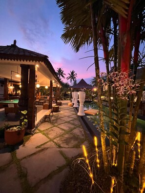 A pool area with a lounge, palm trees, and a sunset sky. - Villa Padi Yogyakarta (Cangkringan)