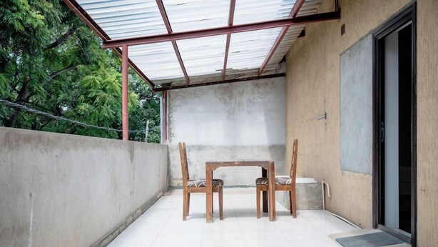 Een overdekt buitengebied met een tafel en stoelen, een betonnen muur en een deur die naar een gebouw leidt.