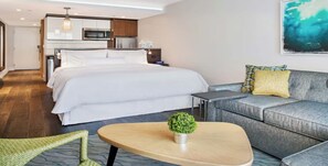 Habitación de hotel con cama, sofá, una mesita y una kitchenette al fondo.