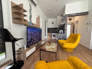 A modern living room with a flat-screen TV, a black chair, a yellow armchair, and a small kitchen area with a washing machine.