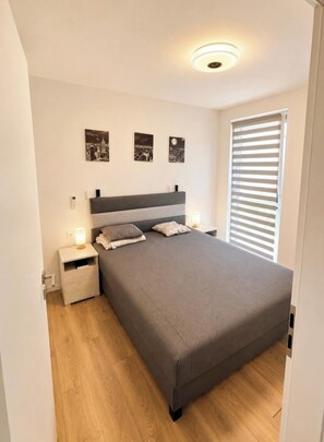 A bedroom with a bed, bedside tables, a nightstand, a wall-mounted light, and a window with blinds.