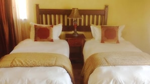 Quarto de hotel com duas camas de solteiro, cabeceira de madeira, mesa de cabeceira com abajur e janela com cortinas.