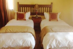 A hotel room with two single beds, a wooden headboard, a nightstand with a lamp, and a window with curtains. - Fox and Squirrel (Dullstroom)