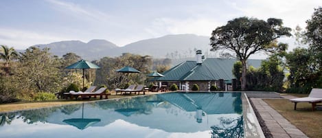 A clear swimming pool with lounge chairs and umbrellas, surrounded by lush greenery and a mountain backdrop.