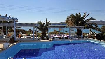 A pool with clear blue water, surrounded by lounge chairs and umbrellas, with a view of the sea and mountains in the background.
