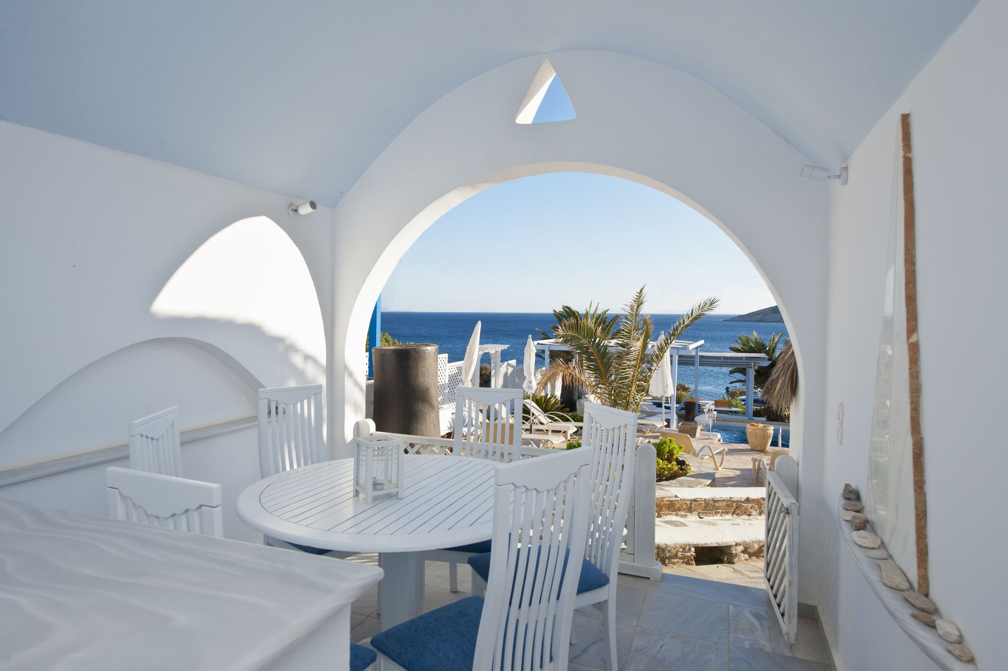 A white outdoor seating area with a table and chairs, overlooking a pool and the sea.
