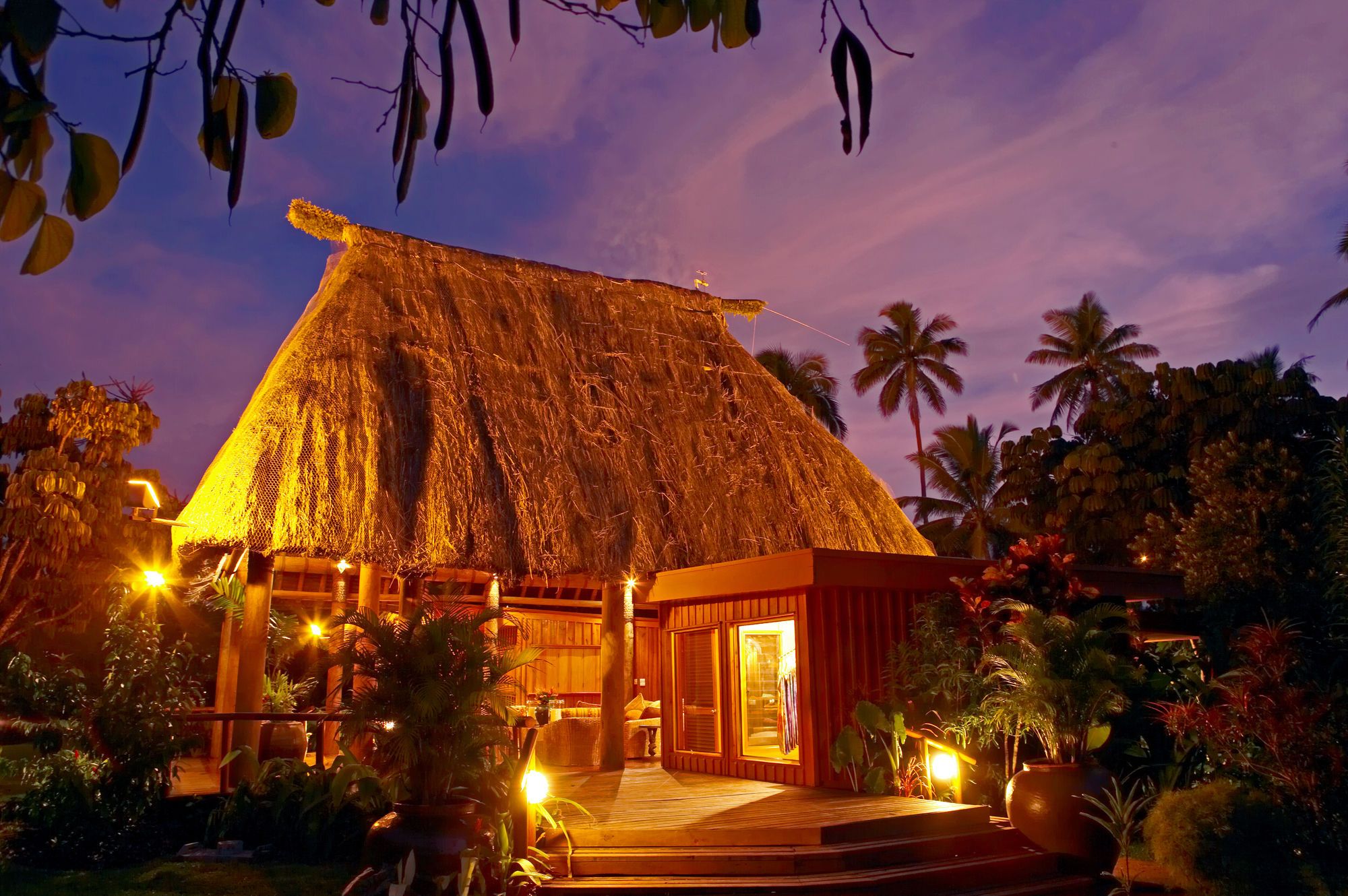 Un bâtiment à toit de chaume, doté d’une véranda en bois, entouré d’une végétation tropicale.