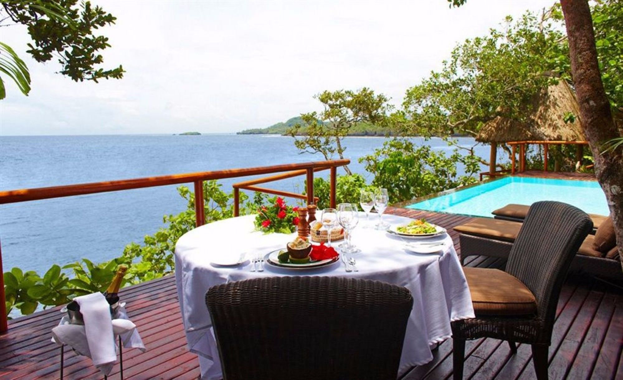 Une table à manger dressée avec des assiettes, des verres et une bouteille, donnant sur une piscine et la mer.