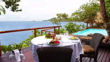 A dining table set with plates, glasses, and a bottle, overlooking a pool and the sea.