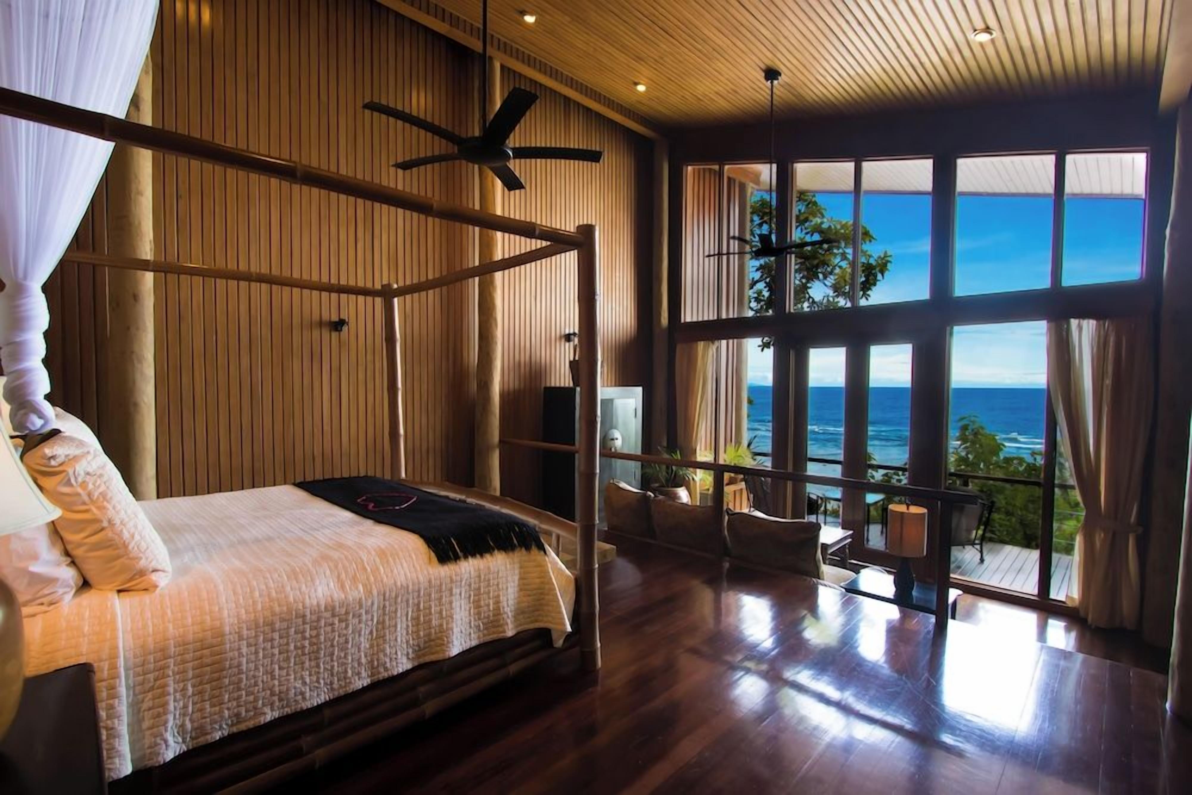 Une chambre avec un lit, un ventilateur de plafond et une grande fenêtre donnant sur l’océan.