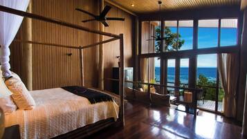 A room with a bed, a ceiling fan, and a large window overlooking the ocean.