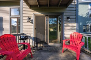 Eine Veranda mit zwei roten Adirondack-Stühlen, einem Grill und einer Glastür zu einem Garten.
