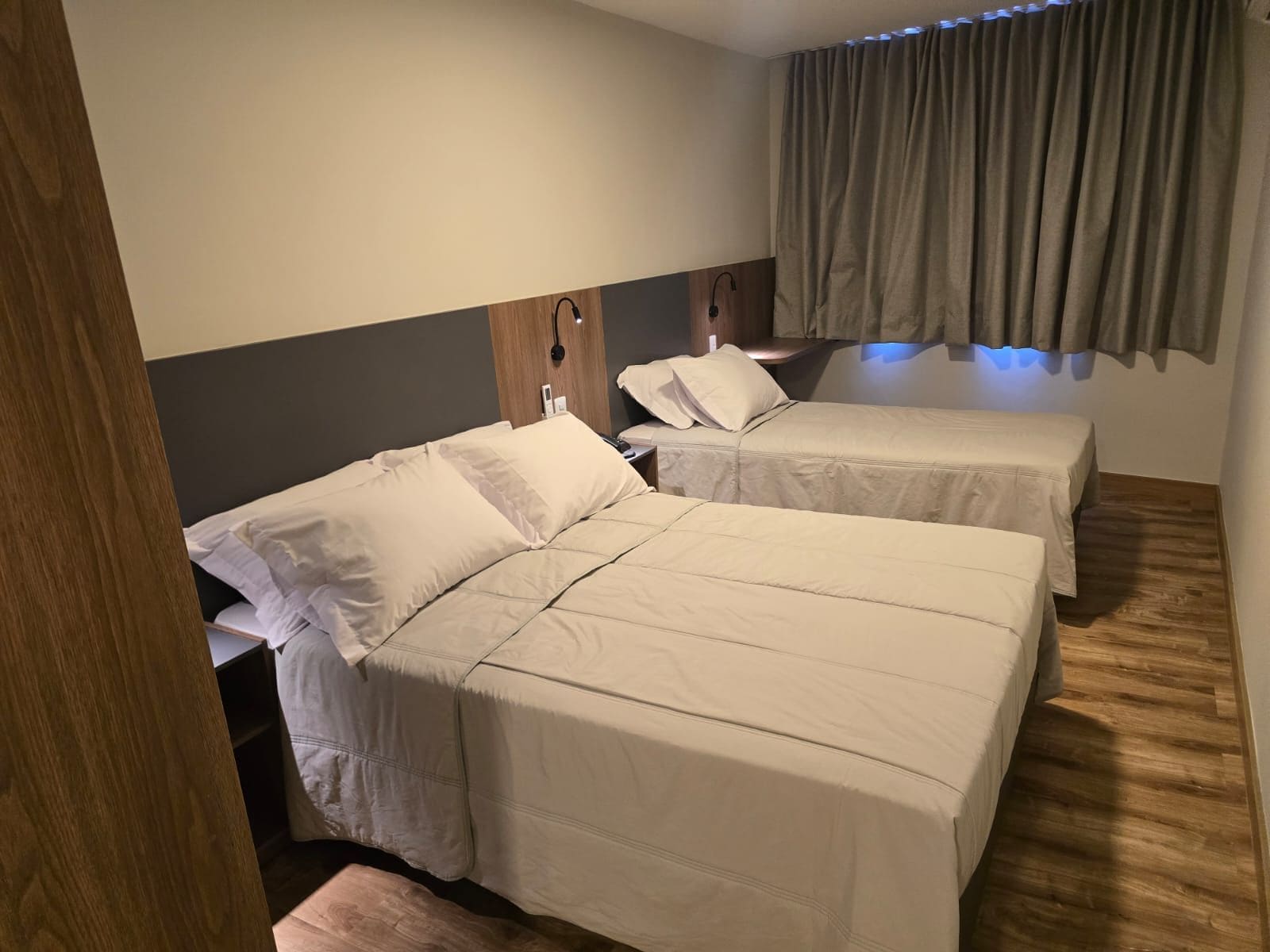 A hotel room with two single beds, a wooden headboard, a bedside table, a nightstand, and a window with curtains.