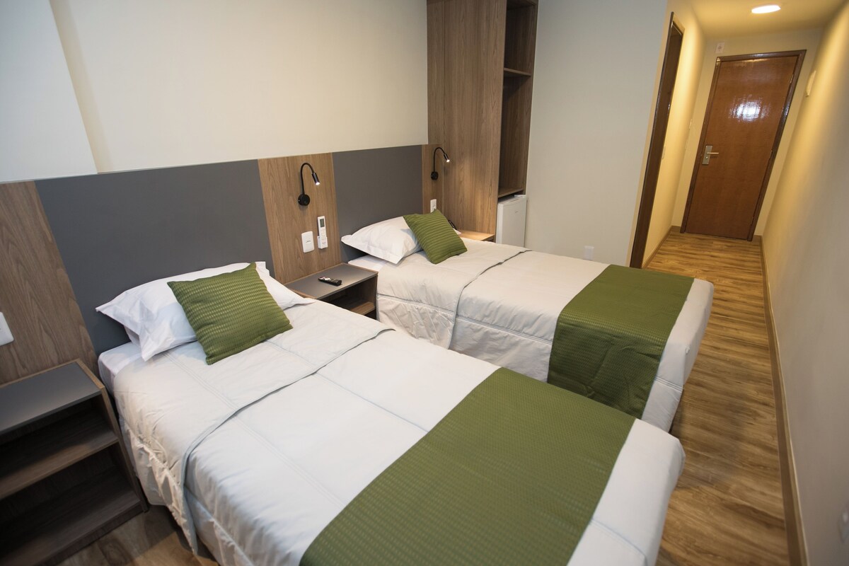 Une chambre d’hôtel avec deux lits, chacun recouvert d’une couvre-lit verte et blanche, une table de chevet, une lampe fixée au mur et une armoire en bois.