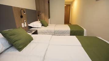 A hotel room with two beds, each with a green blanket and white bedding. There is a nightstand with a remote control and a wall-mounted lamp.
