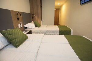 Habitación de hotel con dos camas, cada una con una manta verde y ropa de cama blanca. Se observa un mesita de noche con un control remoto y una lámpara fijada a la pared.