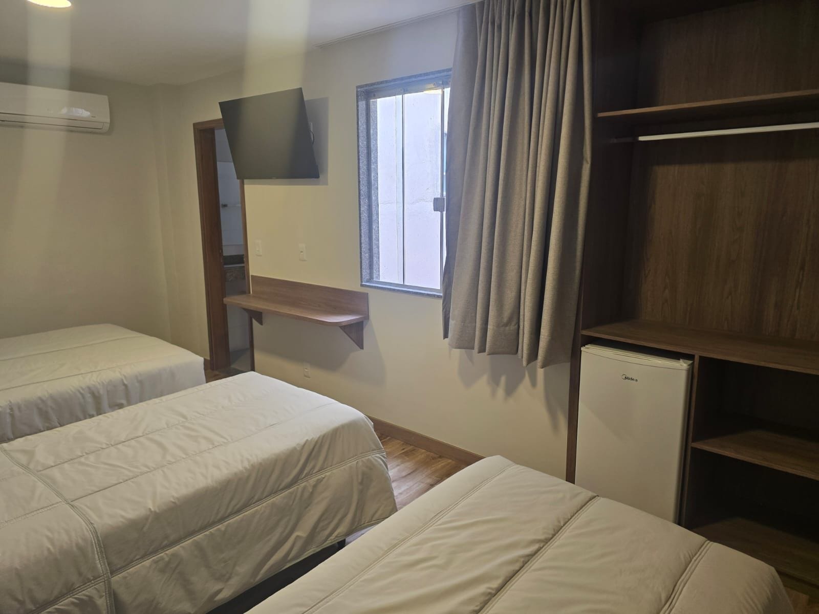 Habitación de hotel con dos camas, un refrigerador pequeño, un espejo, una ventana con cortinas y estantes empotrados.