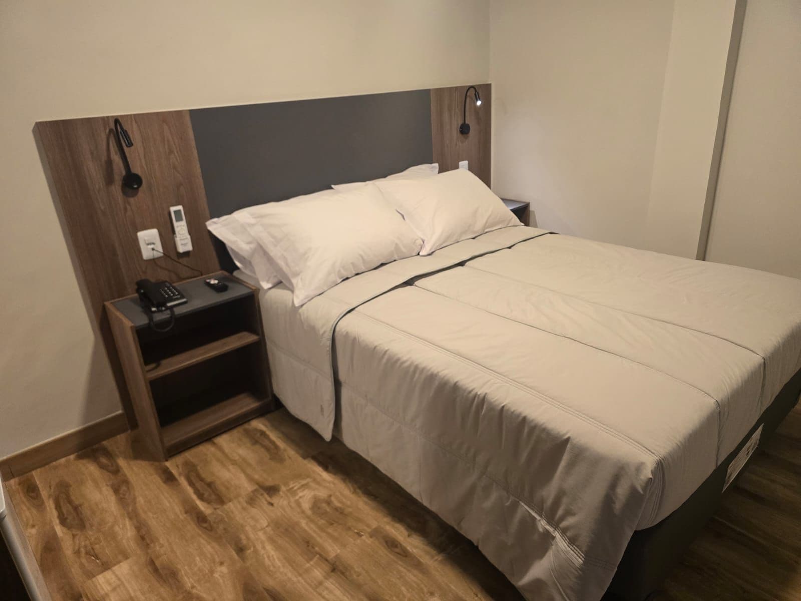 Una cama individual con cabecera, una mesita de noche con un teléfono y una lámpara, y suelo de madera.