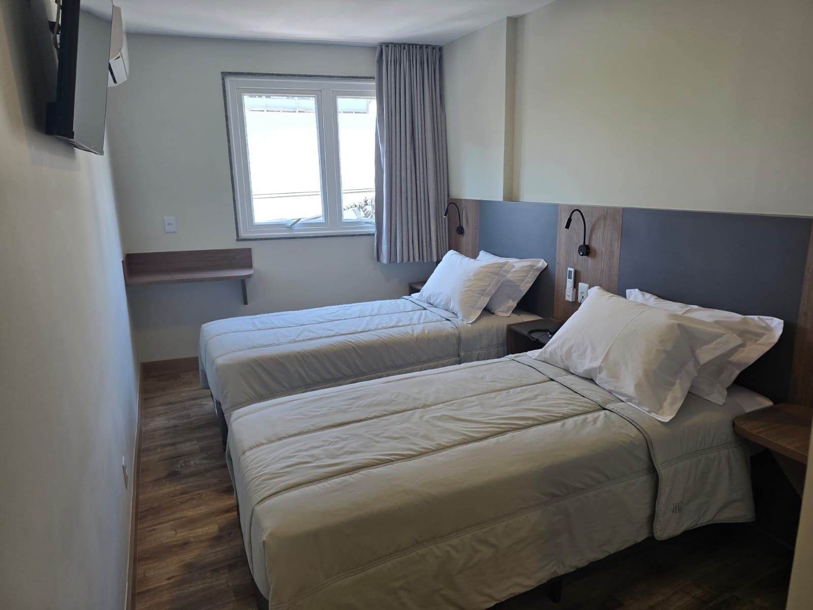 A hotel room with two beds, a window with curtains, a bedside table with a phone, and a wall-mounted TV.