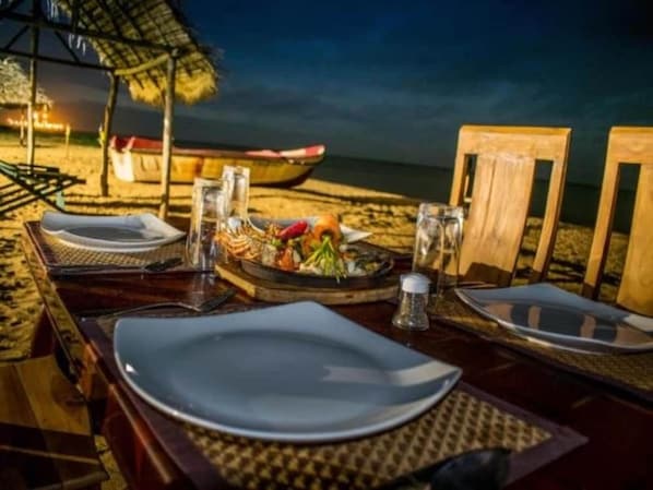 Une table dressée pour deux, avec assiettes, couverts et un verre d’eau, installée sur la plage pour un dîner en bord de mer.
