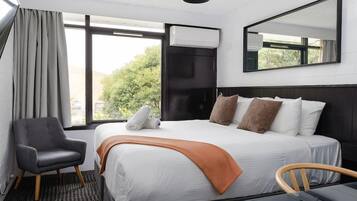 A hotel room with a bed, a chair, a table, a window, an air conditioning unit, and a mirror.