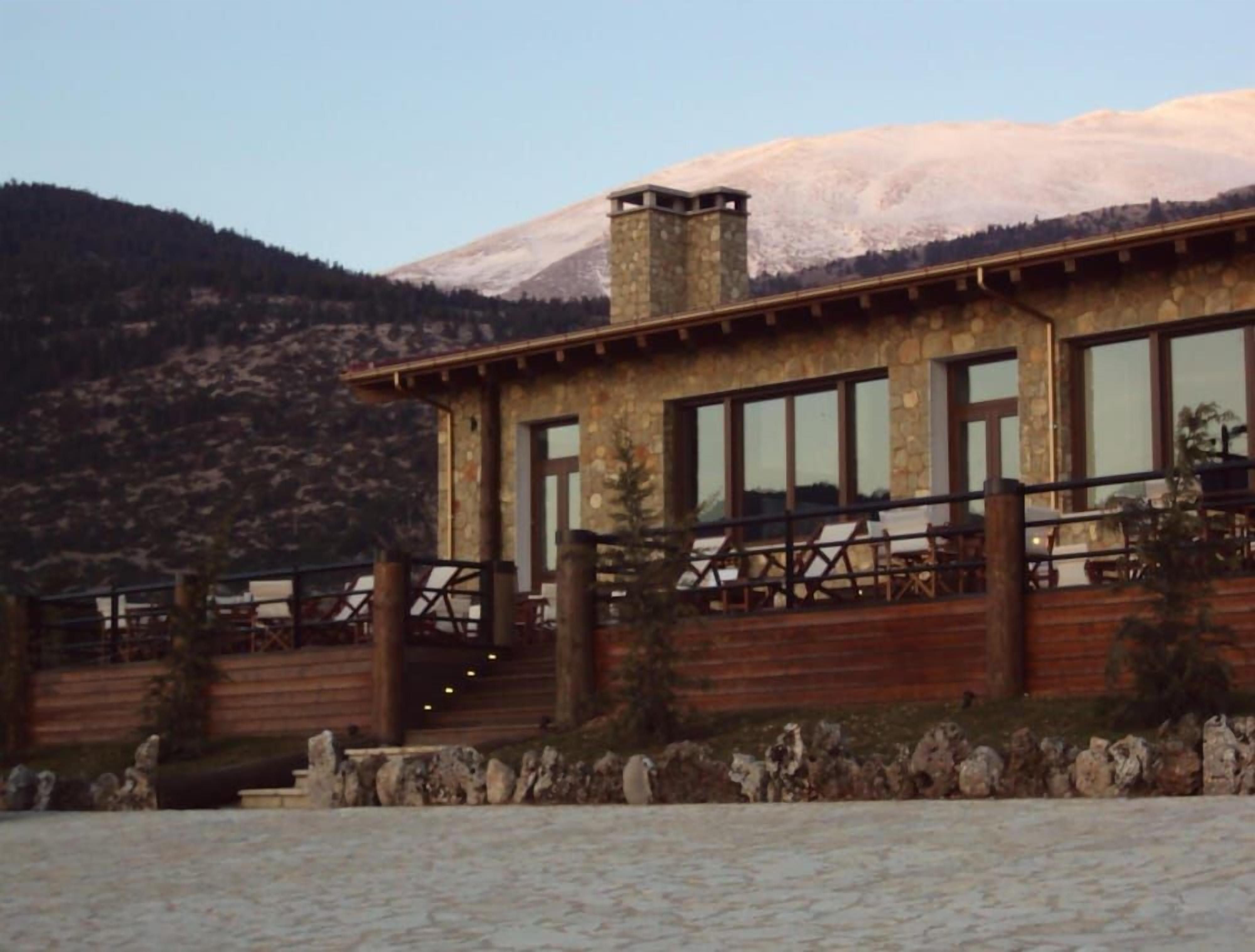 Un edificio de piedra con amplias ventanas y una terraza de madera, con montañas al fondo.