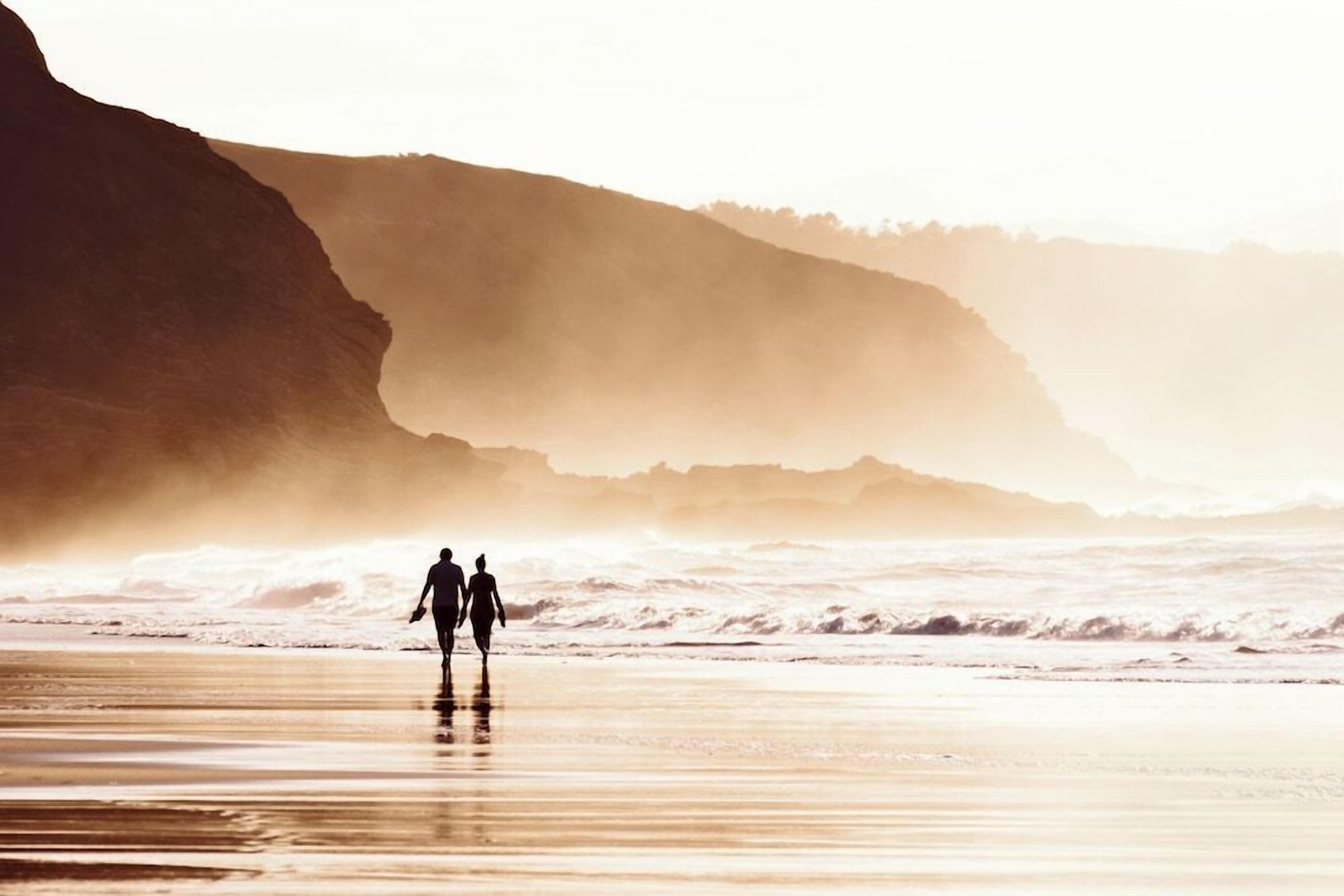 Une scène de plage où l’on voit deux personnes se promener le long du rivage, avec des falaises en arrière-plan et des vagues qui déferlent sur la plage.