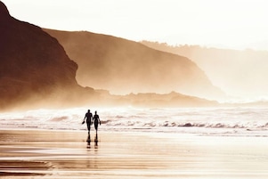 A beach scene with two people walking along the shore, waves crashing in the background, and a cliff on the left.