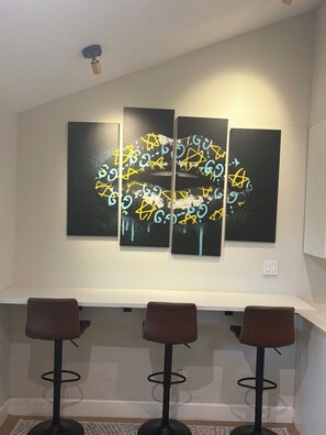 Ein modernes Interieur mit weißer Decke, drei schwarzen Kunstwerken mit gelben Akzenten und einer weißen Theke mit drei braunen Barhocker.