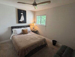 Ein Schlafzimmer mit einem Bett, einem Nachttisch, einer Lampe, einem Deckenventilator, einem Fenster und einem gerahmten Bild an der Wand.