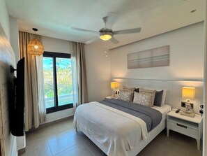 Une chambre à coucher comprenant un lit, des tables de chevet, un ventilateur de plafond, une fenêtre avec des rideaux et une œuvre d’art accrochée au mur.
