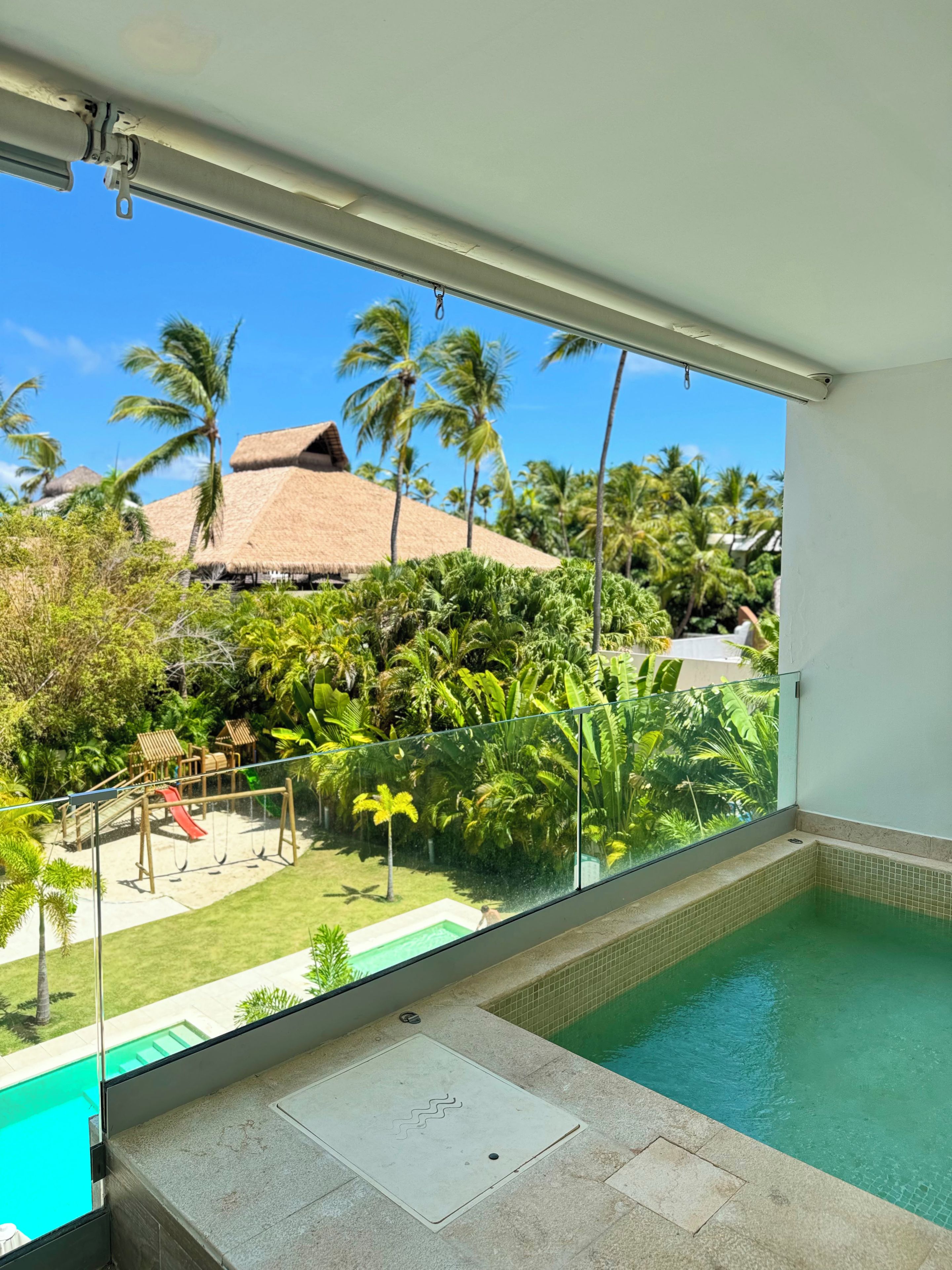 Varanda com guarda-corpo de vidro, vista para uma piscina e um jardim tropical.