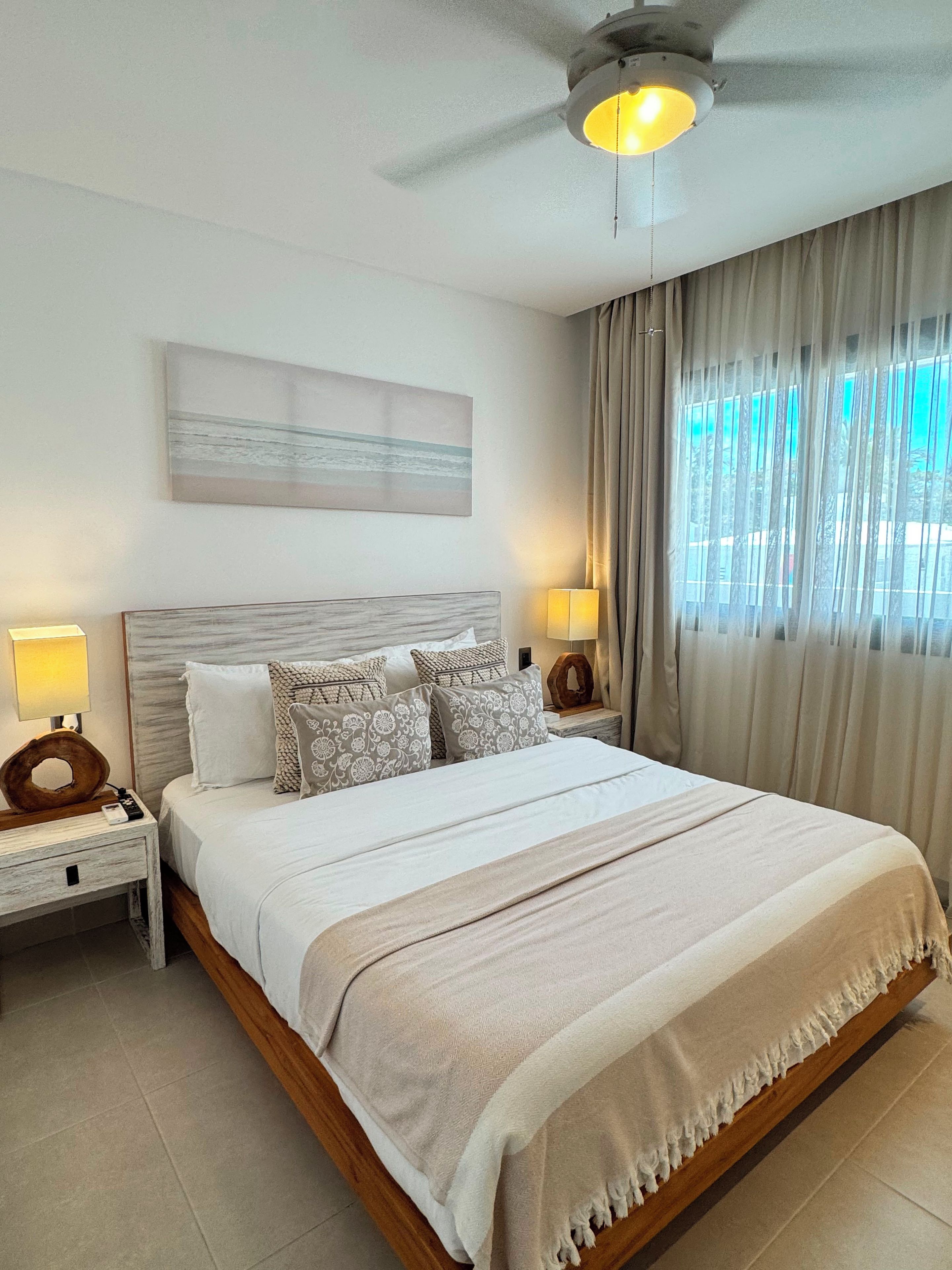Quarto com cama, mesas de cabeceira, ventilador de teto, um quadro e uma janela com cortinas.
