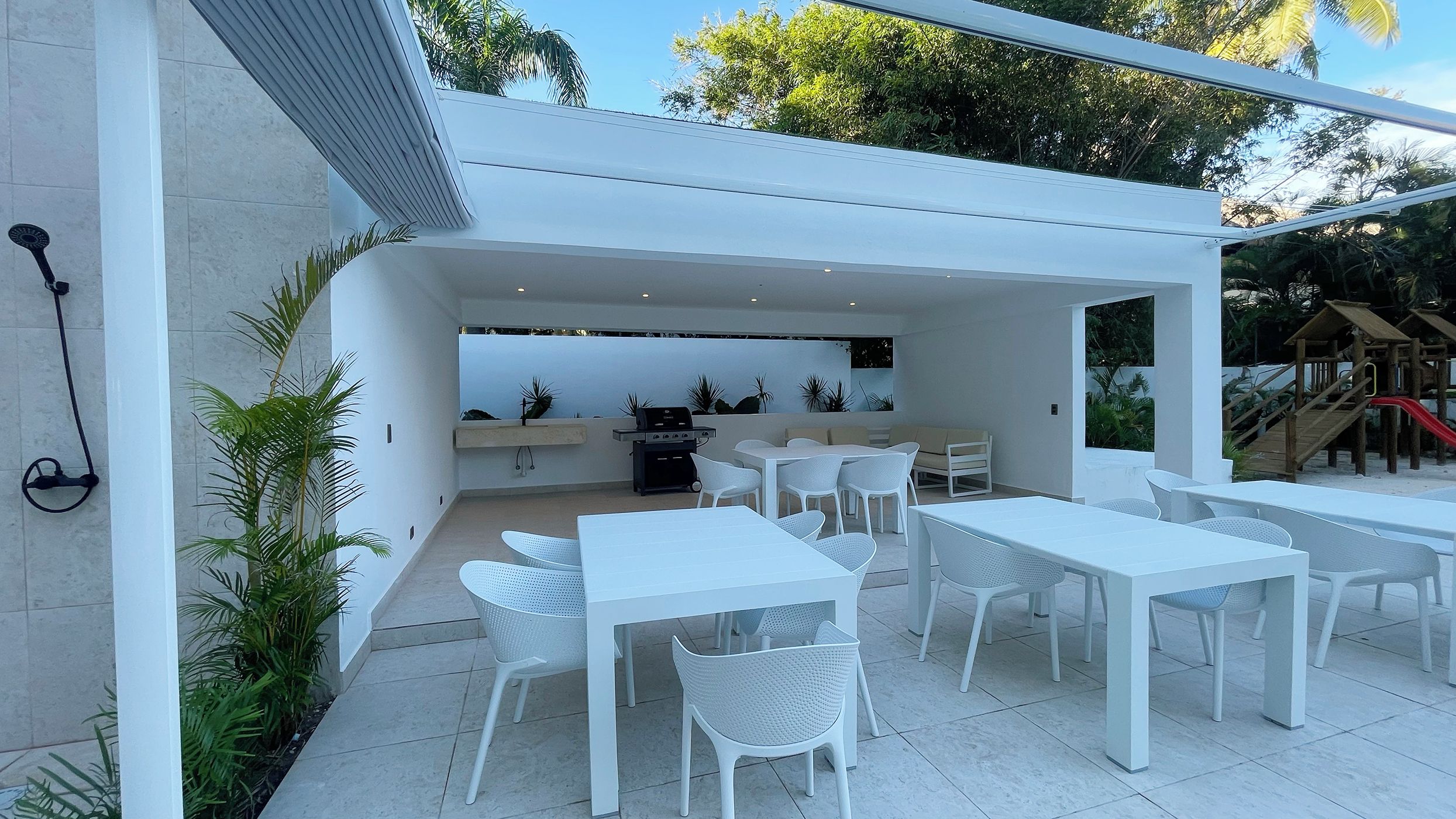 Um pátio externo com mesas e cadeiras brancas, churrasqueira e uma área de recreação infantil ao fundo.