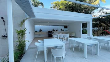 Um pátio externo com mesas e cadeiras brancas, churrasqueira e uma área de recreação infantil ao fundo.