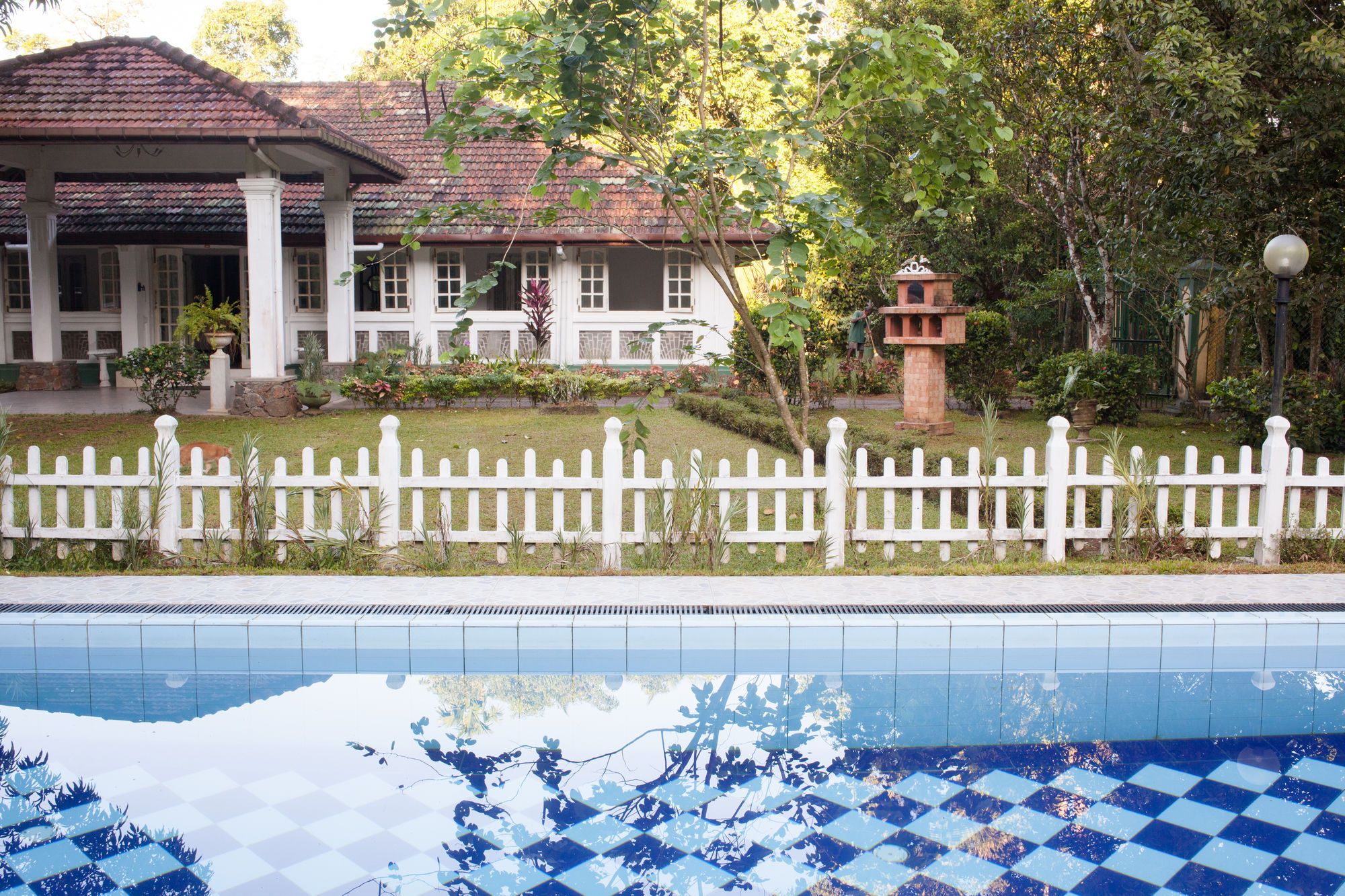 A pool area with a white fence, a building with a tiled roof, and a garden with various plants.