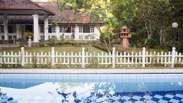 A pool area with a white fence, a building with a tiled roof, and a garden with various plants.
