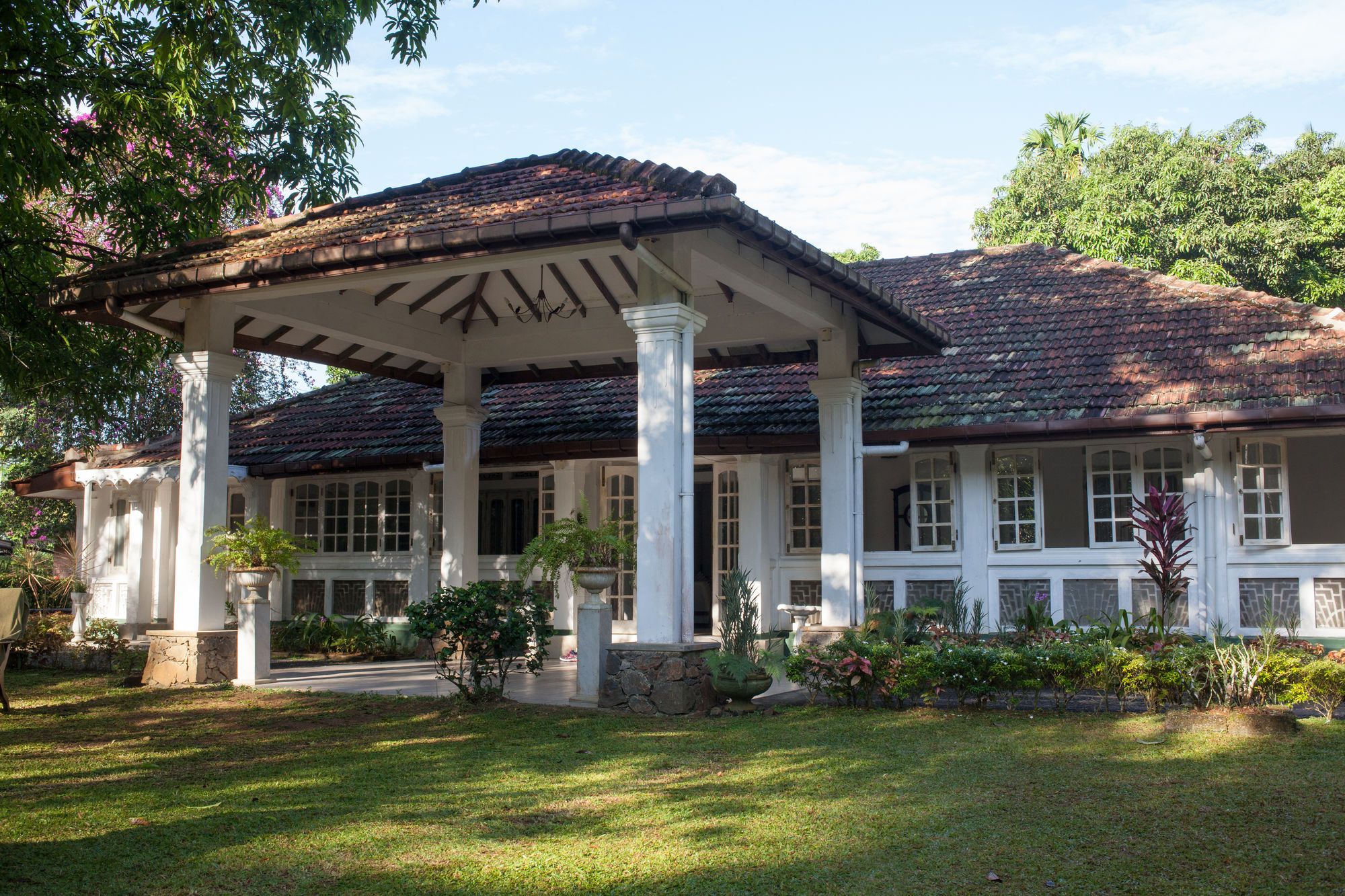 A building with a covered porch supported by columns, a tiled roof, and a well-maintained garden.