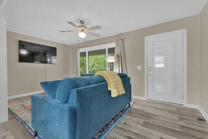 A living room with a blue sofa, a ceiling fan, a television mounted on the wall, and a door with a window. - Newly Renovated, "Daisy" 3 Bedroom, 2 Bath Home! (Hanceville)