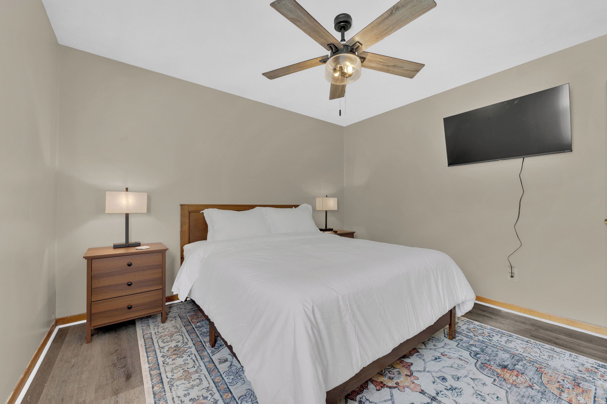 A bedroom with a bed, nightstands, a ceiling fan, a TV mounted on the wall, and a patterned rug.