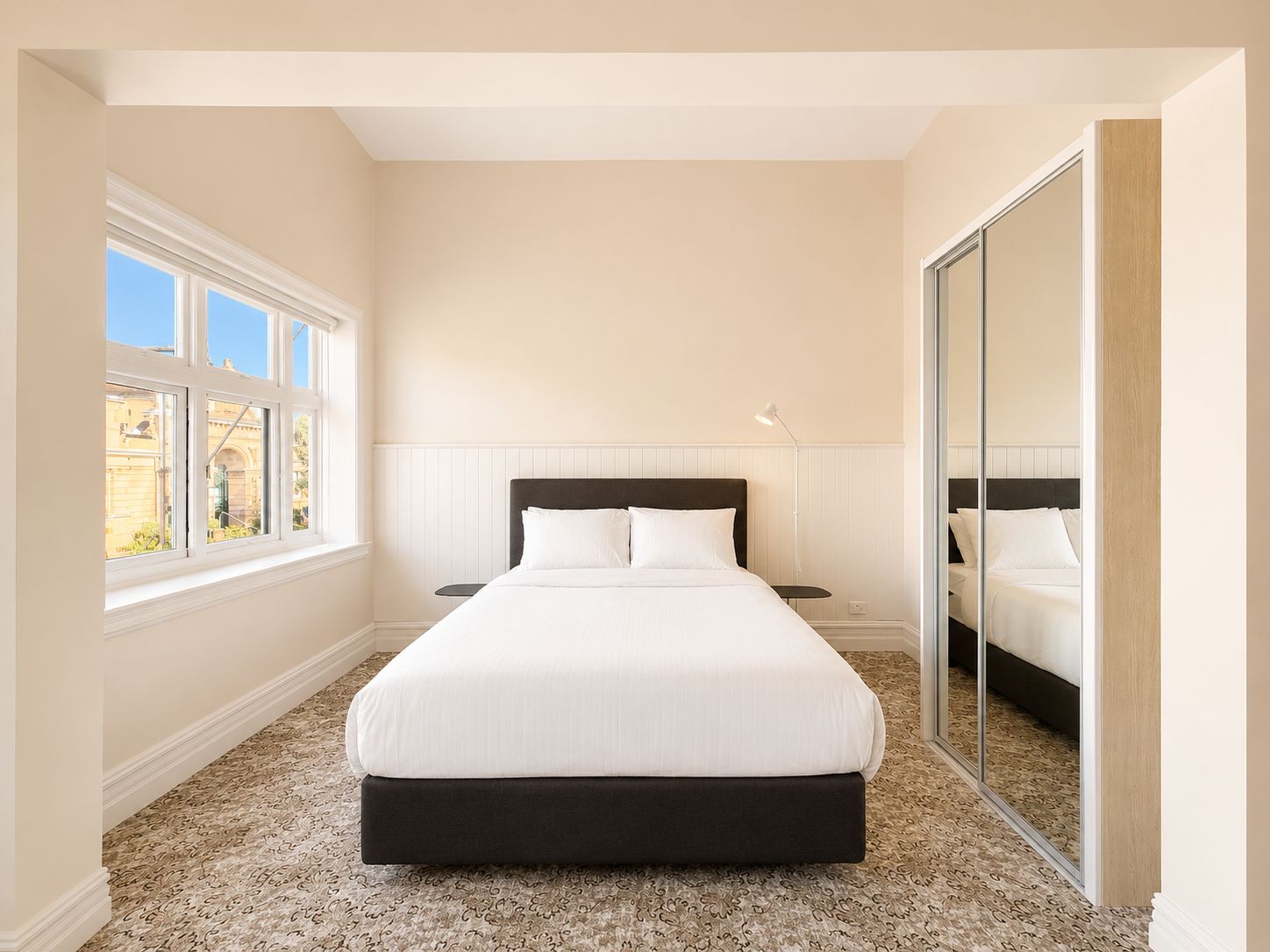 A bedroom with a large bed, a nightstand, a lamp, a window with multiple panes, and a mirror.